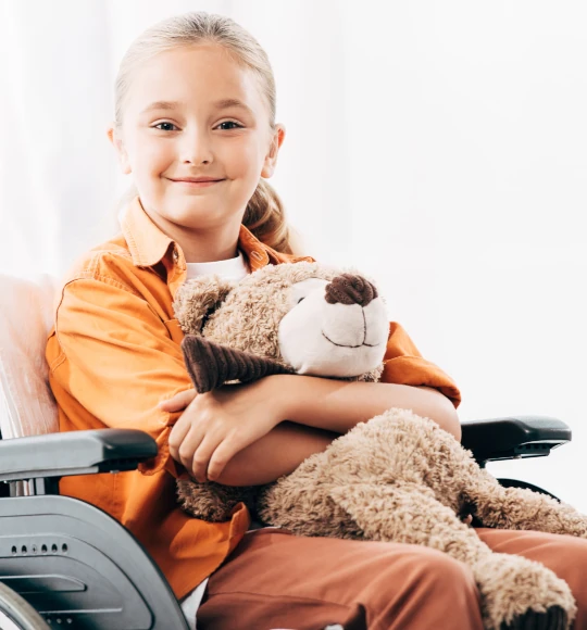 Ein pflegebedürftiges Kind im Rollstuhl mit einem Teddybär im Arm